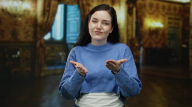 Mavi kazaklı kadın zarif bir sanat galerisinde süslü dekorlu ve dramatik ışıklandırmalı bir şekilde poz vererek büyüleyici ifadesini ve şık arkaplanını vurguluyor..