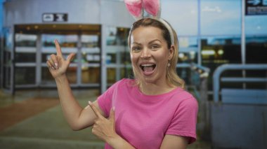 Pembe gömlekli ve tavşan kulaklı bir kadın havaalanı terminalinin girişini işaret ediyor. Heyecan verici..