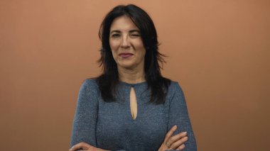 Orta yaşlı, kolları çapraz bir stüdyoda kahverengi duvarlara göz kırpan İspanyol bir kadın..