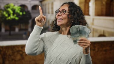 Üniversitede elinde Endonezya Rupiah banknotları tutan bir kadın gülümseyen ve yukarıyı gösteren, güneşli bir günde beyaz kazak ve gözlük takan orta yaşlı bir kadın..