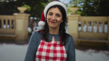 Parkta Noel Baba şapkası takan ve kırmızı önlüklü bir kadın yeşil ağaçların ve taş korkulukların arasında gülümsüyor; bayram neşesi şenliği ruhu.