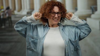 Şehir caddesinde parmaklarıyla kulaklarını kapatan, gürültüden rahatsız olan, sıradan bir kot ve beyaz gömlek giyen, açık hava ortamında tepki gösteren bir kadın..