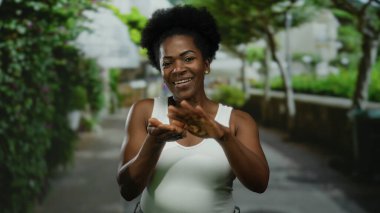 Parkta beyaz atlet giyen bir kadın, elleri ağaçların altında gülümseyen bir jest yapıyor..