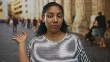 Kadın, eski şehir caddesinde taş sütunların yanında el kaldırmış kameraya bakıyor, kayıtsızlık.