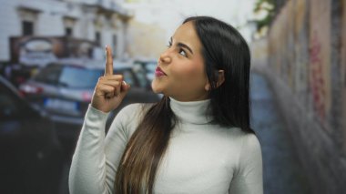 Beyaz kazaklı, uzun saçlı, gülümseyen genç bir kadın park etmiş arabalar ve renkli grafitilerle dolu kalabalık bir şehir sokağında dikilirken....