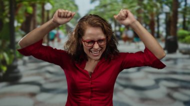 Parlak bir park ortamında neşeli bir kutlama yapan, parlak kırmızı bir gömlek ve gözlük takan, açık havada mutluluk ve heyecanı ifade eden bir kadın..