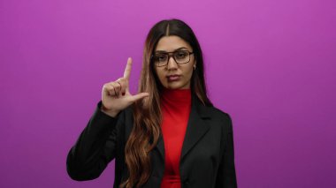 Gözlüklü genç İspanyol kadın pembe arka planda ezik jestler yapıyor, kırmızı ve siyah elbiseler giyiyor, ciddi bir yüz ifadesi sergiliyor..