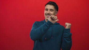 Deniz mavisi kapüşonlu genç İspanyol bir adam canlı kırmızı bir arka plan üzerinde başparmaklarıyla hareket ediyor, pozitifliğini ve güvenini ifade ediyor..