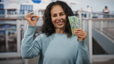 Orta yaşlı bir kadın açık deniz limanı ortamında Avustralya dolarıyla meditasyon yapıyor. Huzuru ve refahı ifade ediyor, elinde banknotlar tutuyor ve memnuniyeti işaret ediyor..
