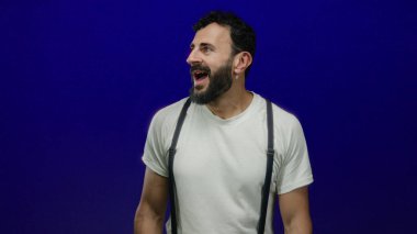 Beyaz tişörtlü ve pantolon askısı takmış İspanyol bir adam dramatik bir etki yaratmak için mavi arka planda sürpriz yapıyor.