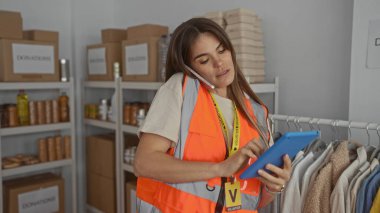 Bağış merkezinde telefonla konuşurken tablet kullanan, arka planda turuncu yelek giyen bir kadın.