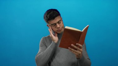 Gözlüklü genç adam, izole edilmiş mavi bir arka plana karşı düşünceli bir ifadeyle kitap okuyor. Modern bir ortamda odaklanma ve konsantrasyon gösteriyor..
