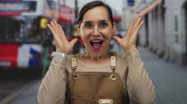 Yoğun caddedeki kırmızı otobüslü önlüklü kadın neşeli duygular ifade ediyor, elleri yüzünü örtüyor sonra sürpriz, şehir ortamında açık havada..