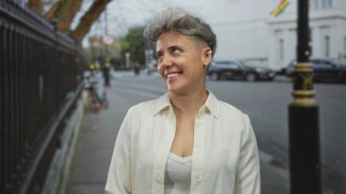 Beyaz tişörtlü, kısa saçlı, yüzü görünen bir caddeye bakan ve arkasında bulanık arabalı bir kadın, iyimserlik.