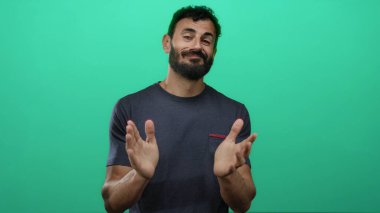 Sakallı İspanyol bir adam canlı yeşil arka planda el çırpıyor, günlük tişört giyiyor, stüdyo ortamında pozitiflik ve dostluk iletiyor..