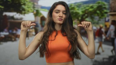 Genç İspanyol kadın, güneşli, yoğun bir caddede, bulanık yayalarla baş parmağıyla göğsünü işaret ediyor..