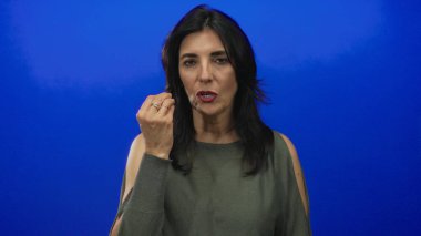 Kadın, canlı mavi stüdyo duvarının önünde hareketli bir el hareketiyle parmak uçlarını çimdikliyor; soru karışıklığı.