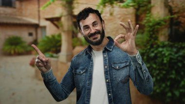 Sakallı İspanyol bir adam yeşil ağaçlarla çevrili güneşli bir parkta jest yapıyor. Kot gömlek giyiyor ve açık havada pozitifliğini gösteriyor..