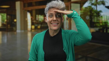 Yeşil hırka giyen bir kadın otel lobisinde el ele tutuşmuş kalkan gözleriyle iç mekanı tarıyor..