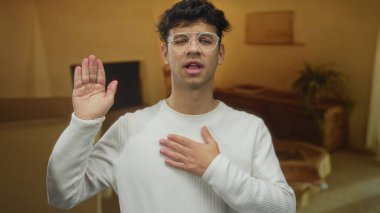 Beyaz gömlekli ve gözlüklü genç adam bir binanın içinde samimi bir konutta elini kaldırır ve göğsüne dokunur; samimiyet.