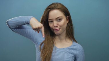 Mavi süveter giyen genç beyaz bir kadın, parlak mavi bir stüdyoda parmağıyla işaret ediyor; neşeli bir eğlence..