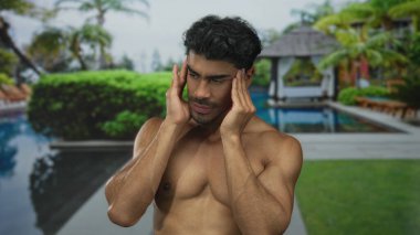 Tişörtsüz adam, güneşli bir sabahta havuz başında tapınak presler; baş ağrısı gerginlik gerginliği.