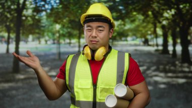 Parkta inşaat işçisi elinde bina düzenini gösteren güvenlik kaskı takan planlar tutuyor.