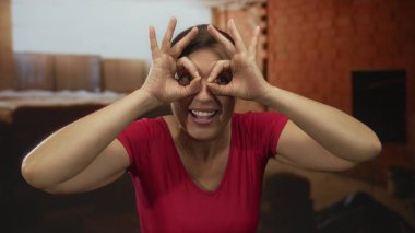 Kırmızı gömlekli bir kadın inşaat alanında gülümserken el dürbünü hareketi yapıyor; merak..