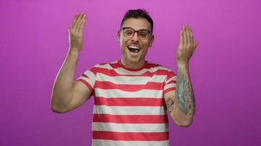 Genç adam neşeli bir şekilde gülümsüyor, elleri kapalı pembe arka planda, çizgili gömlek ve gözlük takıyor.