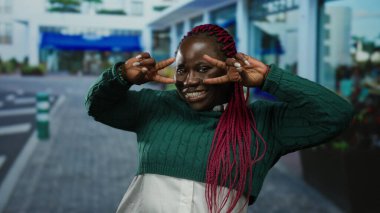 Şehir sokaklarında yeşil süveter ve pembe örgüler takmış, açık hava ortamında neşe ve çeşitliliği ifade eden barış işaretleriyle gülümseyen bir kadın..