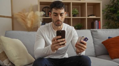 Kredi kartı ve telefonlu genç adam şık oturma odasında koltukta oturuyor, rahat bir evde modern yaşam tarzı ve finansal yönetim sergiliyor..