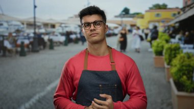 Gözlüklü ve önlüklü genç adam şehir caddesinde açık hava kafeleri ve canlı bir atmosferi olan güvenli bir şekilde duruyor..