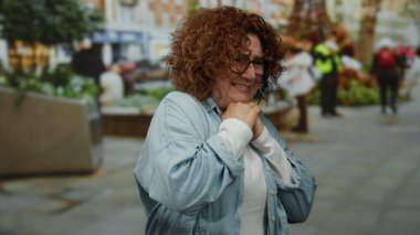 Kıvırcık saçlı bir kadın kalabalık bir şehir caddesinde insanlarla ve yeşillikle, orta yaşlı ve esmer bir şekilde şehir dışında endişeli bir ifadeyle ortaya çıkıyor..