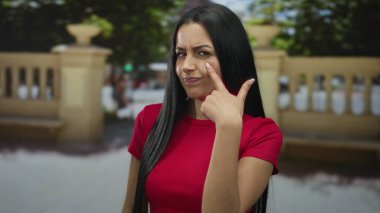 Kırmızı tişörtlü İspanyol kadın ağaçlar ve korkuluklarla dolu bir sokak ortamında şüpheli bir ifade takınıyor..