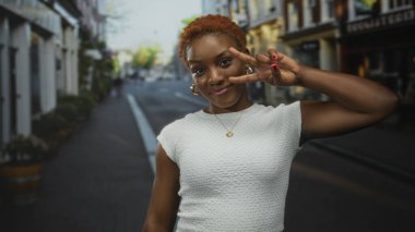 Beyaz desenli bluz ve altın kolye takan kadın şehir sokaklarında barış işareti gösteriyor; neşe ve güven.