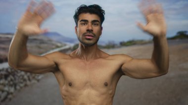 Çıplak göğsü olan adam mavi gökyüzü altında kayalık yol boyunca iki elini kaldırır; açıklık bağlantısını selamlar.