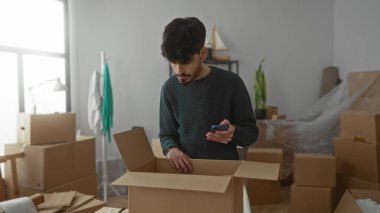 Genç adam yeni bir dairede kutuları açıyor. Elinde akıllı bir telefon var. Etrafı malzemelerle çevrili..