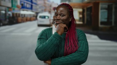 Şehir caddesinde kendine güvenen bir kadın yeşil süveter ve pembe örgüler takıyor. Etrafı şehir hayatı ve araçlarla çevrili. Açık havada güven ve şehir enerjisi taşıyor..