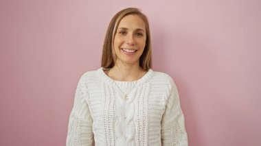 Pembe bir duvarın önünde gülümseyen bir kadın beyaz bir kazak giyiyor sakin ve pozitif bir ifade sergiliyor.