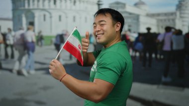 Yeşil tişörtlü genç bir adam arka planda yürüyen insanlarla birlikte Pisa, İtalya 'nın eğimli kulesinin önünde İran bayrağı taşıyor..
