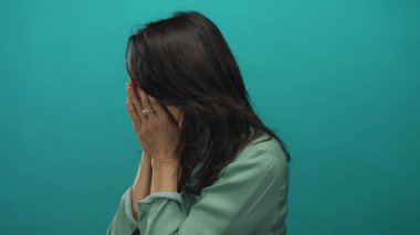 Orta yaşlı, suratını buruşturan ve turkuaz bir stüdyo çekiminde elleriyle yüzünü örten İspanyol kadın..