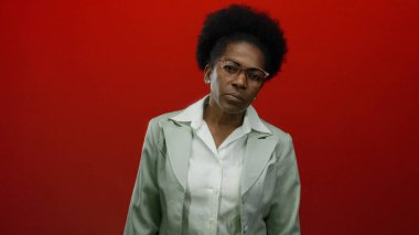 Gözlüklü bir kadın, canlı kırmızı bir arka planda, beyaz bir bluz ve takım elbise giyiyor, düşünceli bir şekilde düşünüyor ve kendine güveniyor..