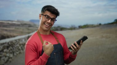 Gözlüklü ve kırmızı kazaklı genç bir adam, elinde tablet, sakin bir caddede gülümsüyor..