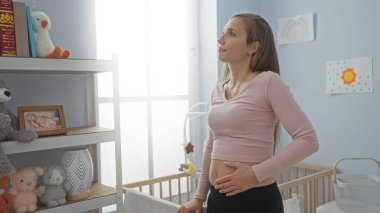 Yatak odasında beşikte duran, karnını nazikçe tutan, bebek oyuncakları ve kitaplarla çevrili bir kadın, evde besleyici ve beklenen bir atmosfer olduğunu gösteriyor..