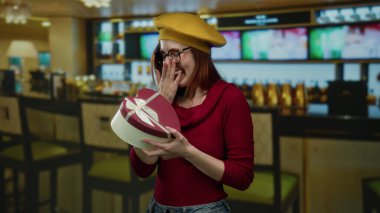 Kalp şeklinde bir hediye kutusu tutan bir kadın, şık bir restoran ortamında, şık bir bere ve gözlük takıyor, sıcak ve davetkar bir ortam yaratıyor..
