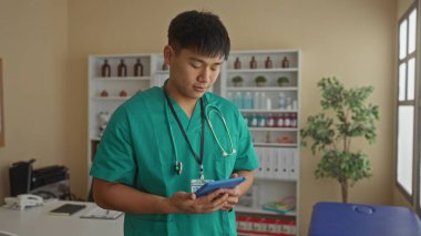 Yeşil önlüklü genç, tıbbi malzeme ve ekipmanlarla dolu bir hastane odasında tablet kullanarak sakin bir sağlık hizmeti ortamı sergiliyor..