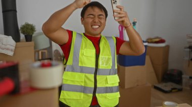 Yansıtıcı yelek giyen Asyalı adam dağınık oturma odasında kutuları taşırken heyecanlı bir şekilde akıllı telefon kullanıyor..