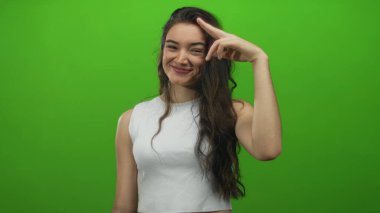 Genç bir kadın, parlak yeşil bir stüdyo ortamında gelişigüzel selamlaşırken geniş bir gülümseme sergiliyor..