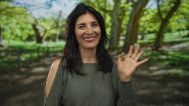 Orta yaşlı bir kadın, güneşli bir ormanda çıplak el sallayarak sıcak bir şekilde gülümserken, sıcak ve samimi bir şekilde selamlıyor..