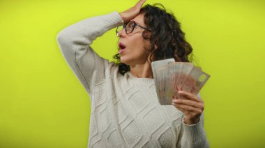 Orta yaşlı bir kadın, izole edilmiş sarı arka planda dolarları tutuyor. Parayla şaşkınlık ve heyecan yaratıyor..